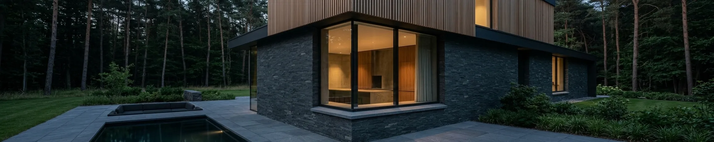 A single-storey house of dark slate and timber at dusk, a warmly lit corner window facing the garden, with a pool to one side and pine woodland behind.