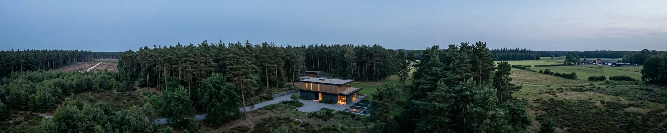 An aerial view of the house set within pine woodland, with open heath and farmland stretching away beyond the treeline.