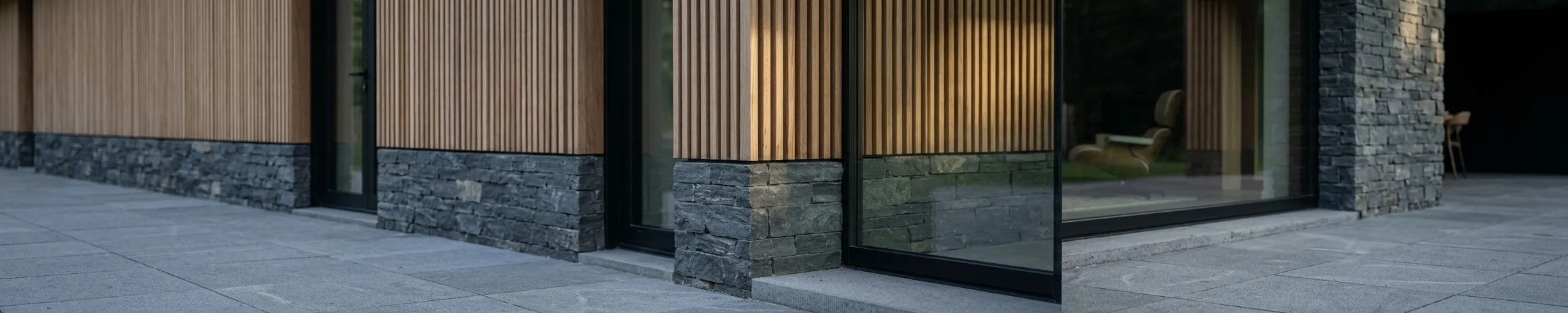 The façade in close view: vertical timber cladding above a dark slate base, with full-height glazing reflecting the trees and a chair visible inside.