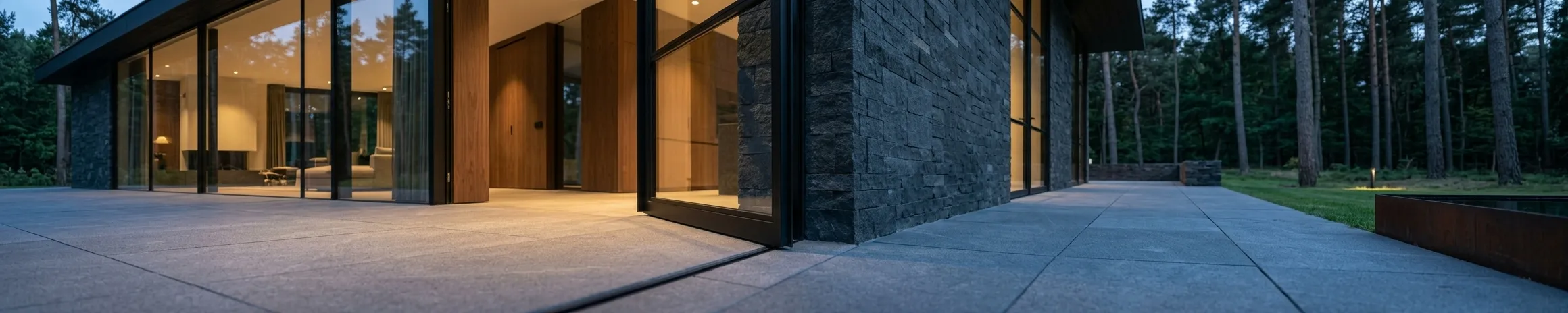The house at dusk under a deep dark eaves line, stone and timber walls meeting broad stone paving, with a weathered steel edge to one side.