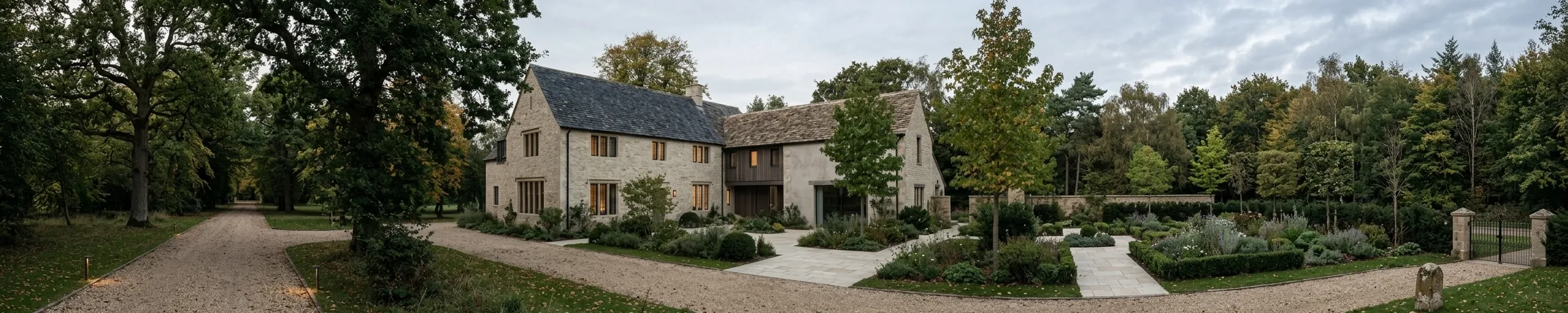 A stone house with a steep slate roof and a lower timber-clad wing, approached by a gravel drive, with clipped hedging, planted borders and mature oaks around it.