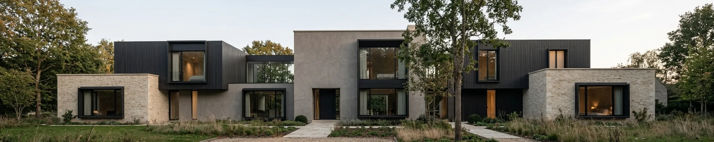A long, low house composed of pale stone, render and dark timber volumes with deep-set black windows, fronted by grasses and a stone path through planting.