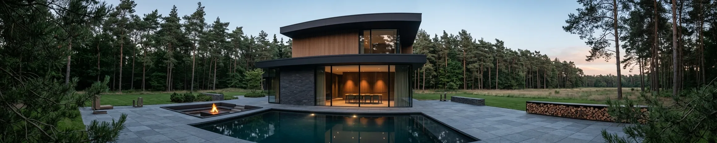 A house with a gently curved roof profile at dusk, its lit interior reflected in a long pool, with a fire bowl and a stacked log store on the stone terrace.