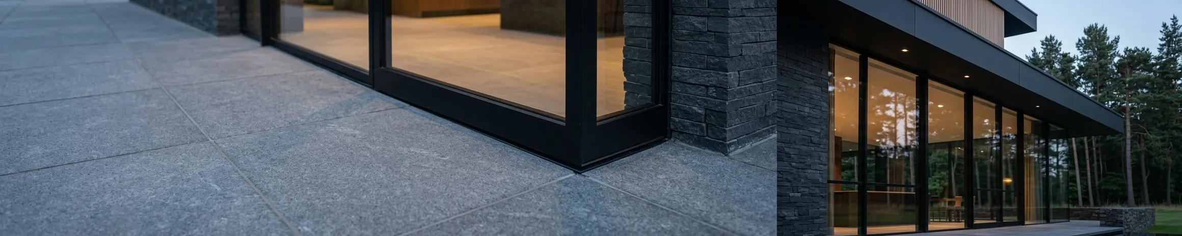 Large stone paving running up to full-height glazing in slim dark frames, with a lit interior visible beyond.