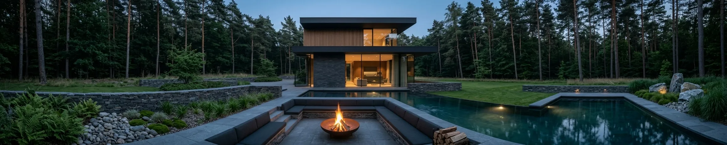 A lit house seen across a still pool at dusk, a fire bowl burning on the sunken terrace between the water and the building.