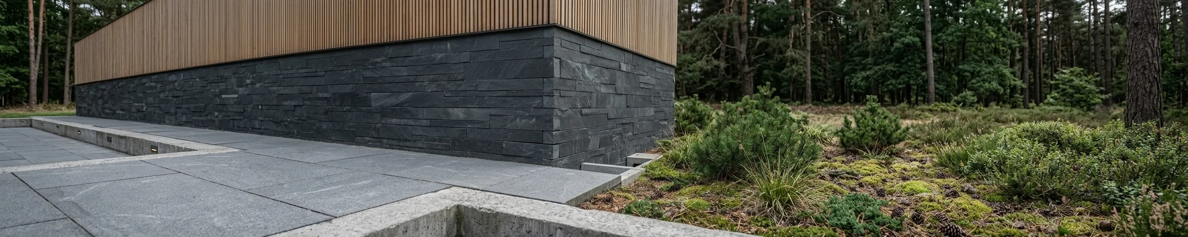 Vertical timber cladding above a dark stone wall, meeting broad stone paving, with heath planting and pines alongside.