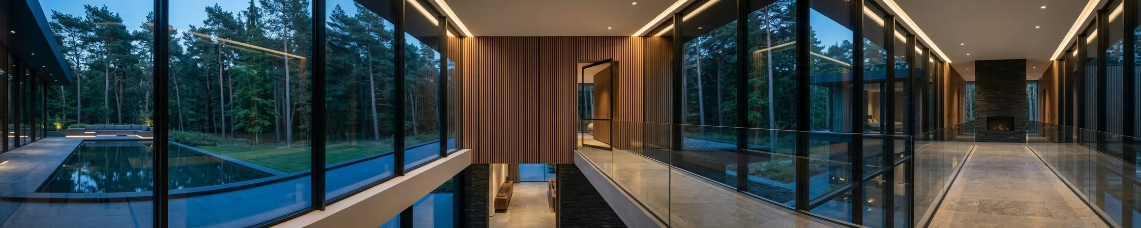 An upper-level gallery with a glass balustrade and a timber-lined wall, full-height glazing looking out over a pool, and a stone fireplace in the room beyond.